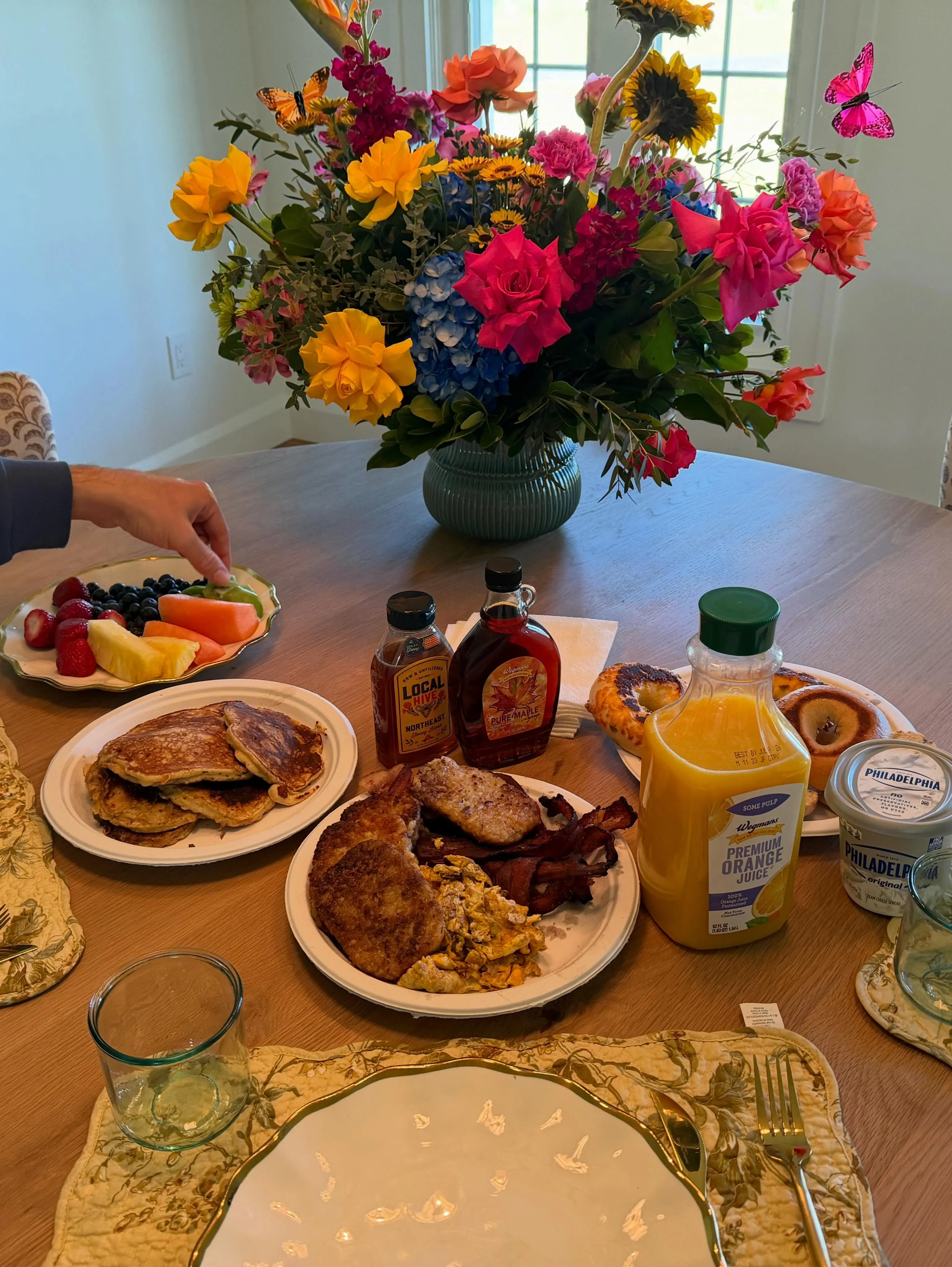 A Mother's Day breakfast with pancakes, eggs, bacon, fruit, orange juice, and a large colorful flower arrangement.