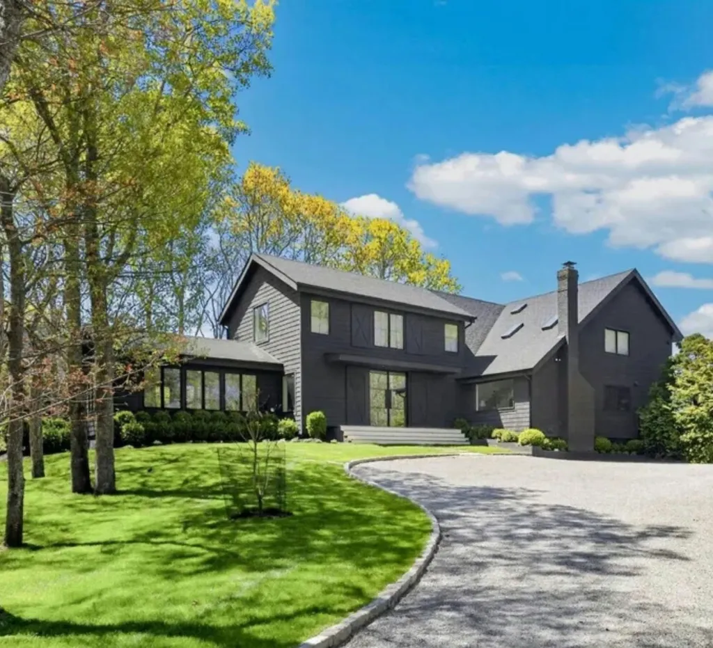 A large, dark gray house with multiple gables and windows, a long gravel driveway, and a manicured lawn surrounded by trees under a bright blue sky.