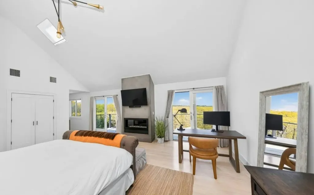 A modern bedroom with a bed in the foreground, a fireplace, a mounted TV, a desk, and doors leading to a balcony overlooking a field.