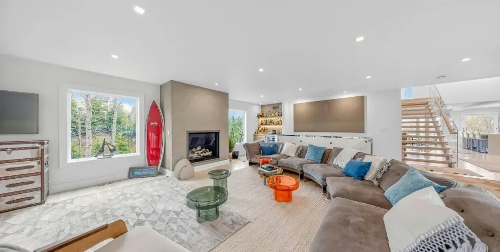 A spacious living room featuring a large gray sectional sofa, a fireplace, a built-in bar, and a modern wooden staircase.