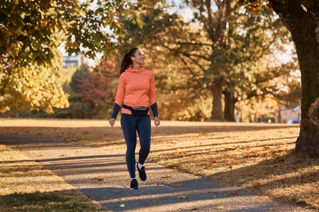 A happy athletic woman with earbuds in an orange hoodie and dark leggings walks through an autumn park.