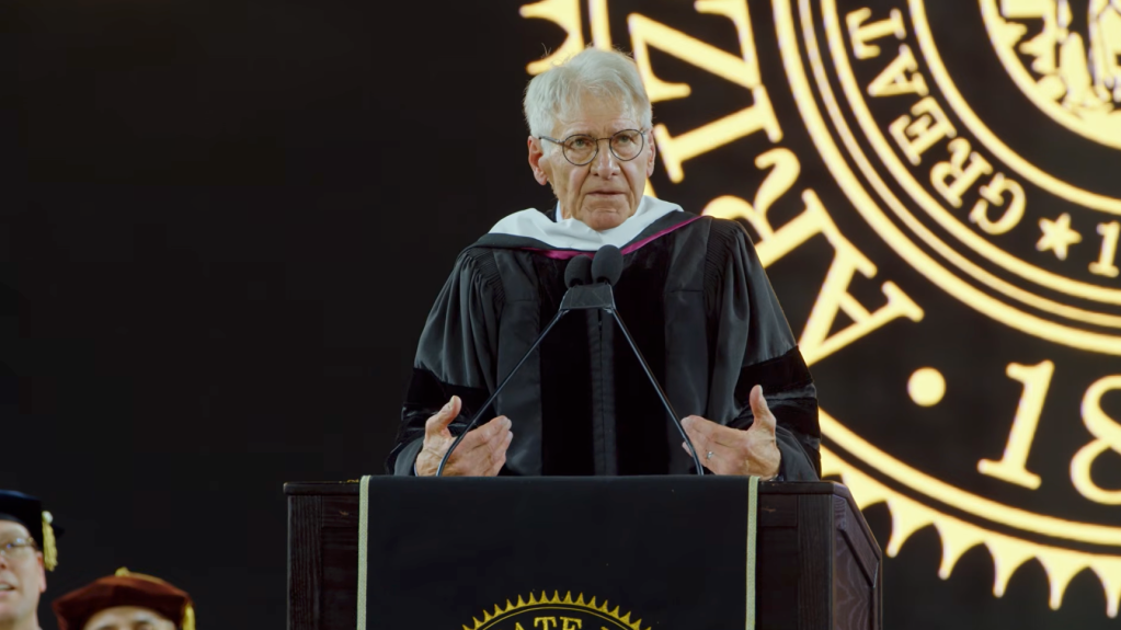 Harrison Ford speaking at a podium in graduation robes with a large golden seal in the background.