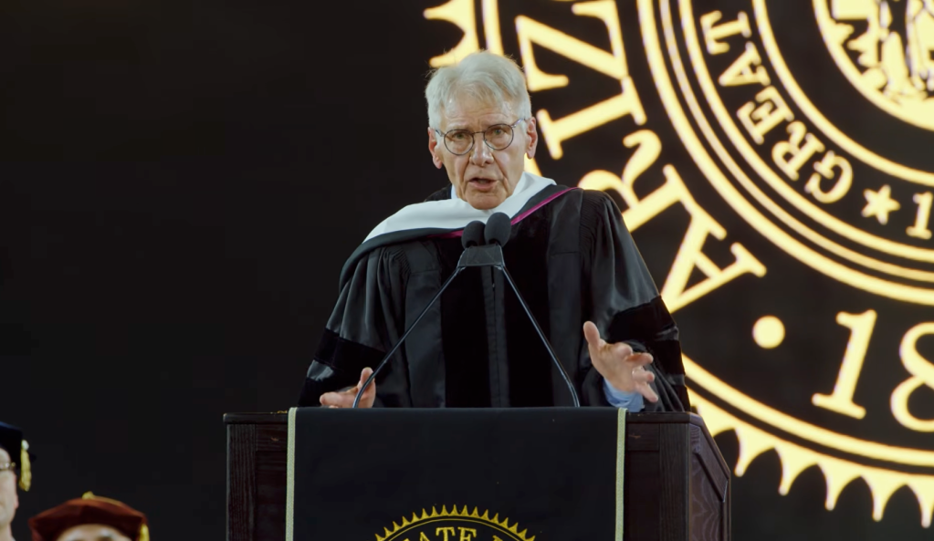Harrison Ford speaking at a podium in a graduation gown, with a large golden seal in the background.