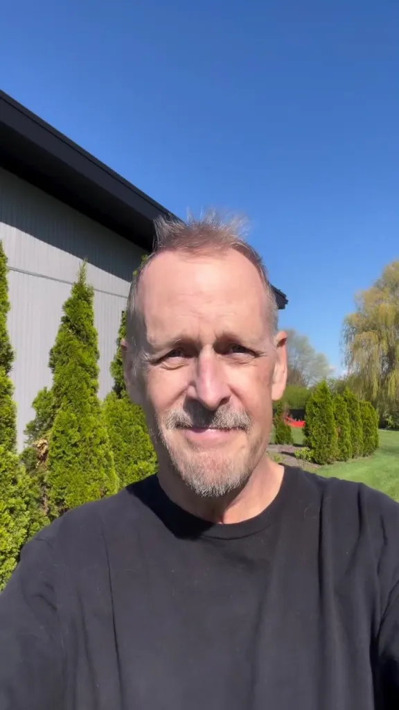 Dave Coulier in a black shirt, outdoors with green trees and a building in the background.