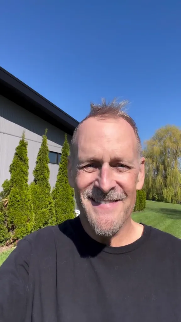 Dave Coulier smiles at the camera in a black t-shirt against a clear blue sky.