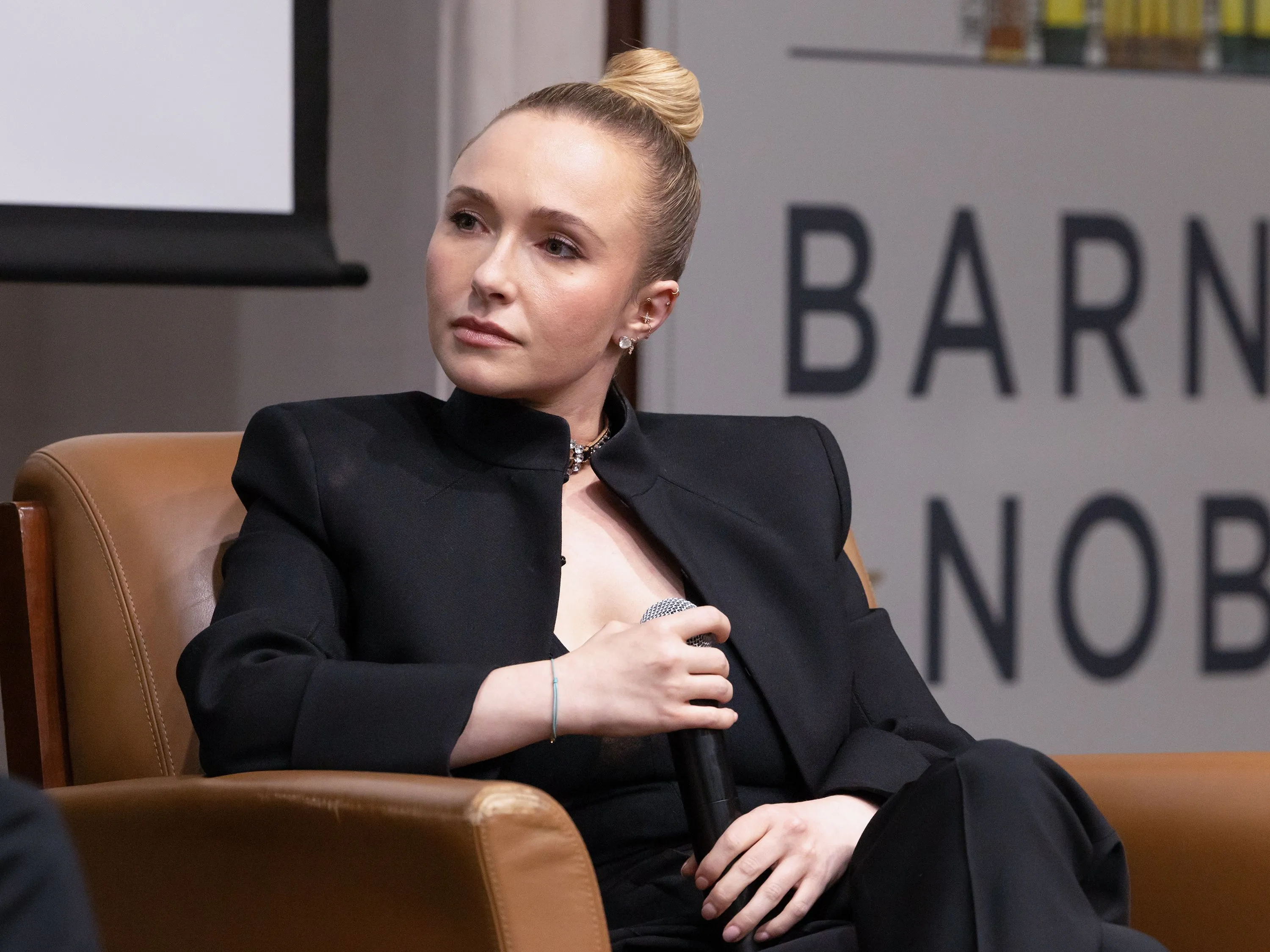 Hayden Panettiere in a black suit, holding a microphone while seated.