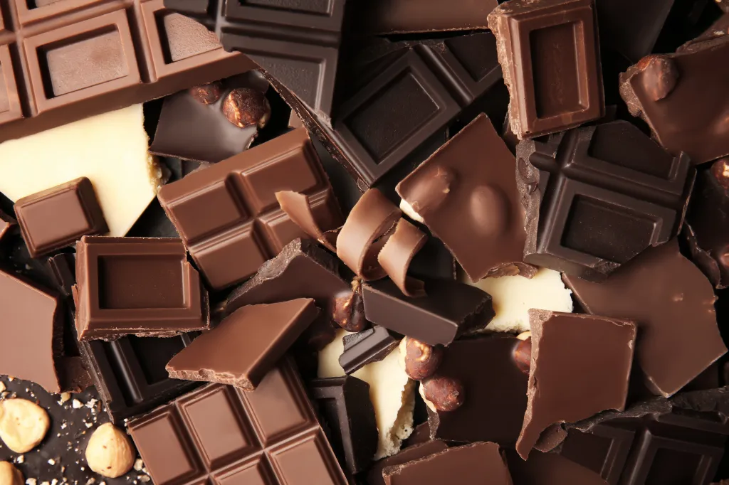 A close-up of a heap of broken chocolate pieces of various types and shades, including milk, dark, and white chocolate, with some hazelnut pieces and chocolate shavings scattered among them.