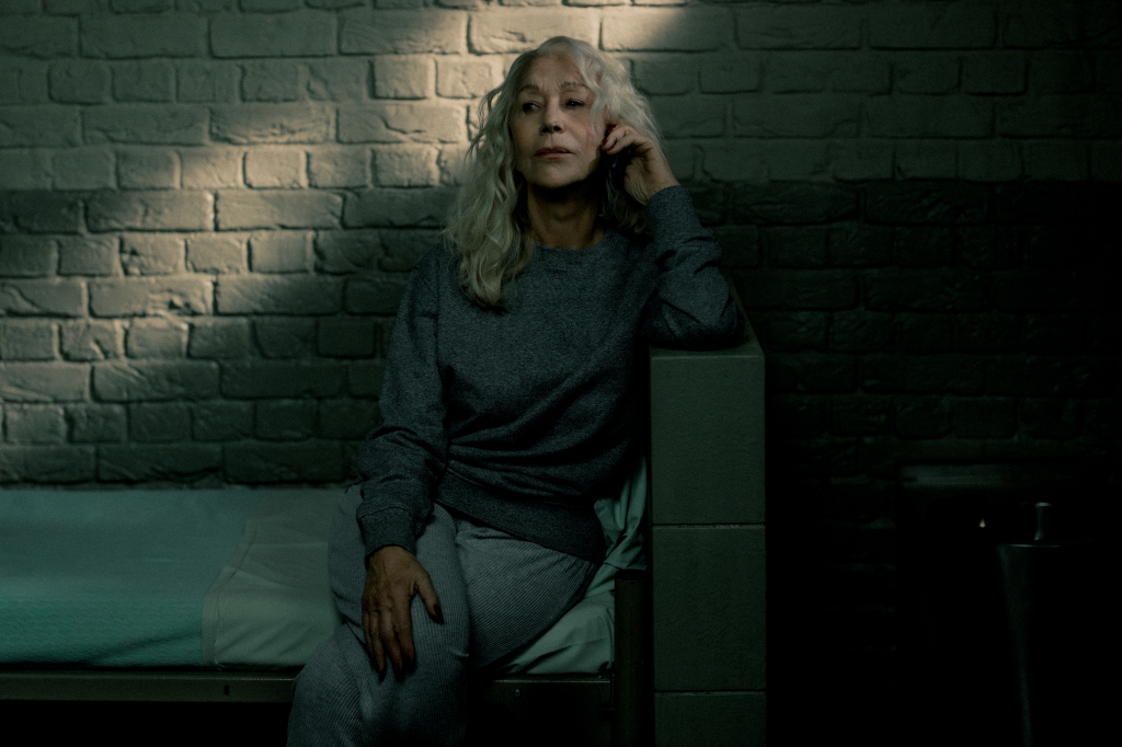 Helen Mirren as Maeve Harrigan sits on a cot in a dark room.