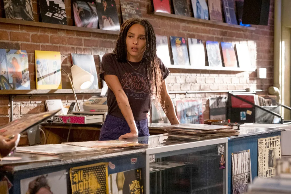 Zoe Kravitz as Rob in a record store, leaning on a counter next to stacks of records and a register.