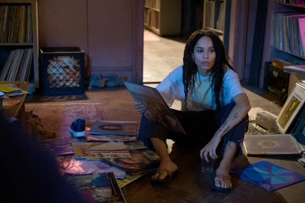 Zoe Kravitz as Rob, holding a record album and sitting on the floor surrounded by records.