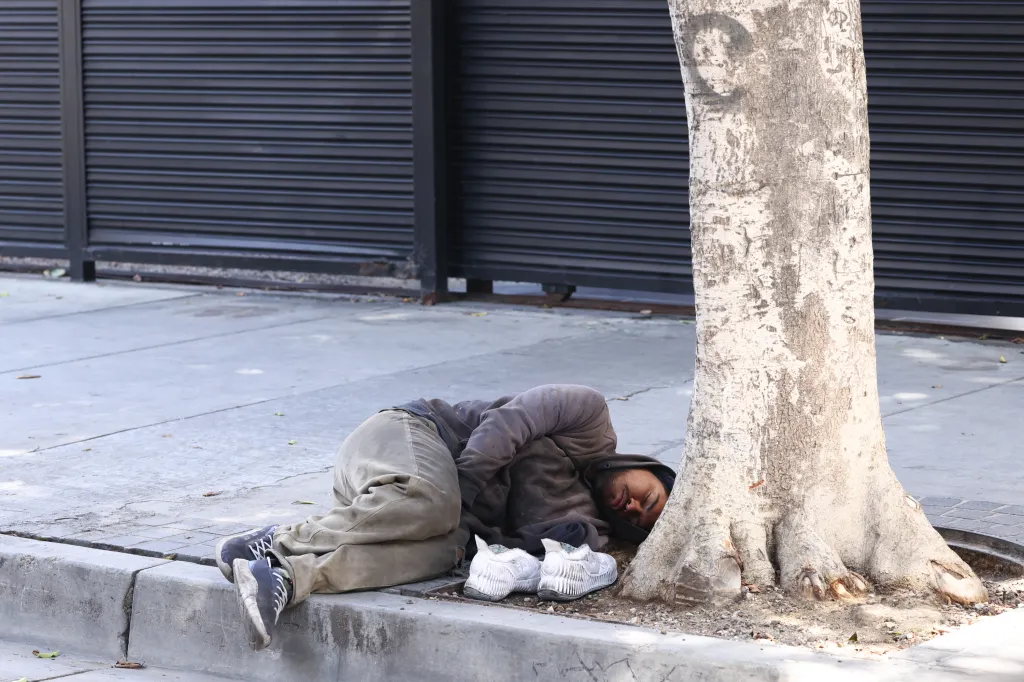 A homeless person sleeping on a sidewalk next to a tree, with a pair of shoes beside them.