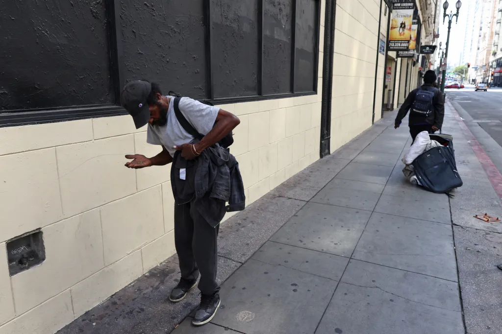 A homeless man on a downtown street looking at his open palm, while another person walks away pushing a rolling suitcase filled with belongings.
