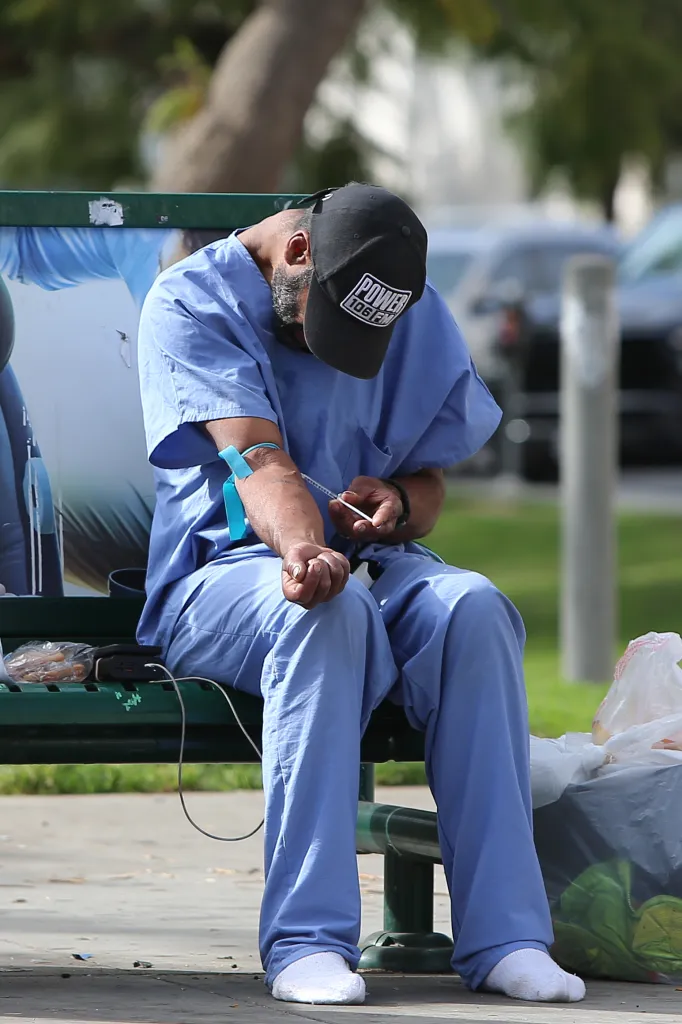 A person in light blue scrubs and a black baseball cap injects a substance into their arm while sitting on a park bench.