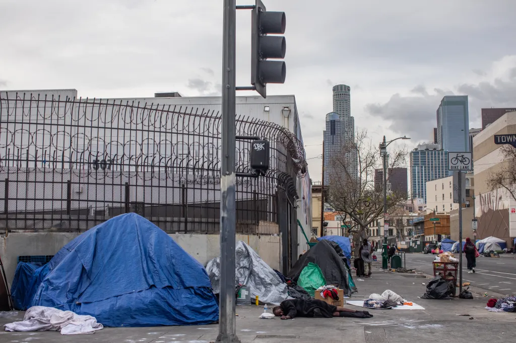 A homeless man sleeps on the ground in front of tents and a metal fence with barbed wire in Los Angeles, with tall buildings in the background.