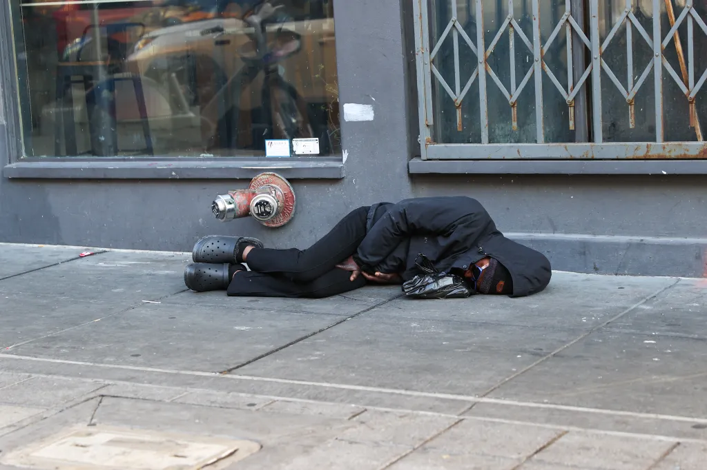 A homeless person in San Francisco sleeps on a sidewalk against a building wall, wearing a black jacket and black Crocs, with a red fire hydrant nearby.