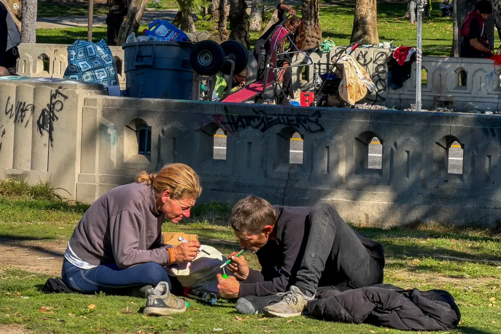 Homeless people resting at MacArthur Park, a man lying on the grass smoking a cigarette and a woman next to him looking at a small box.