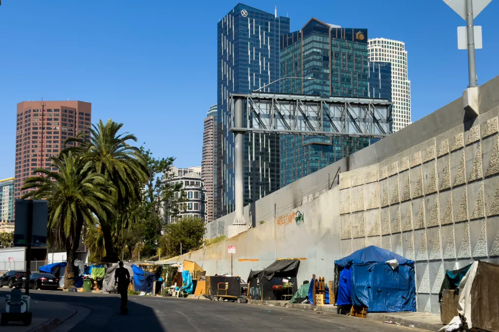 Homeless encampments line a street alongside a concrete wall, with downtown Los Angeles skyscrapers in the background.