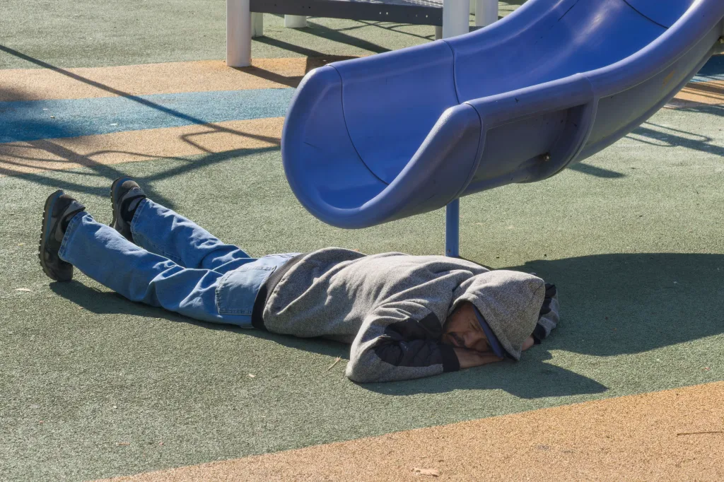 A homeless person sleeping on a green and tan playground surface next to a blue slide.