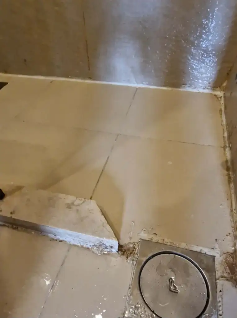 A dirty shower with moldy grout and peeling sealant, with a metal drain cover on the floor.