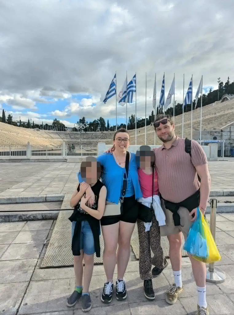 The Edward family smiling in front of the Panathenaic Stadium in Athens, Greece, with Greek flags flying in the background.