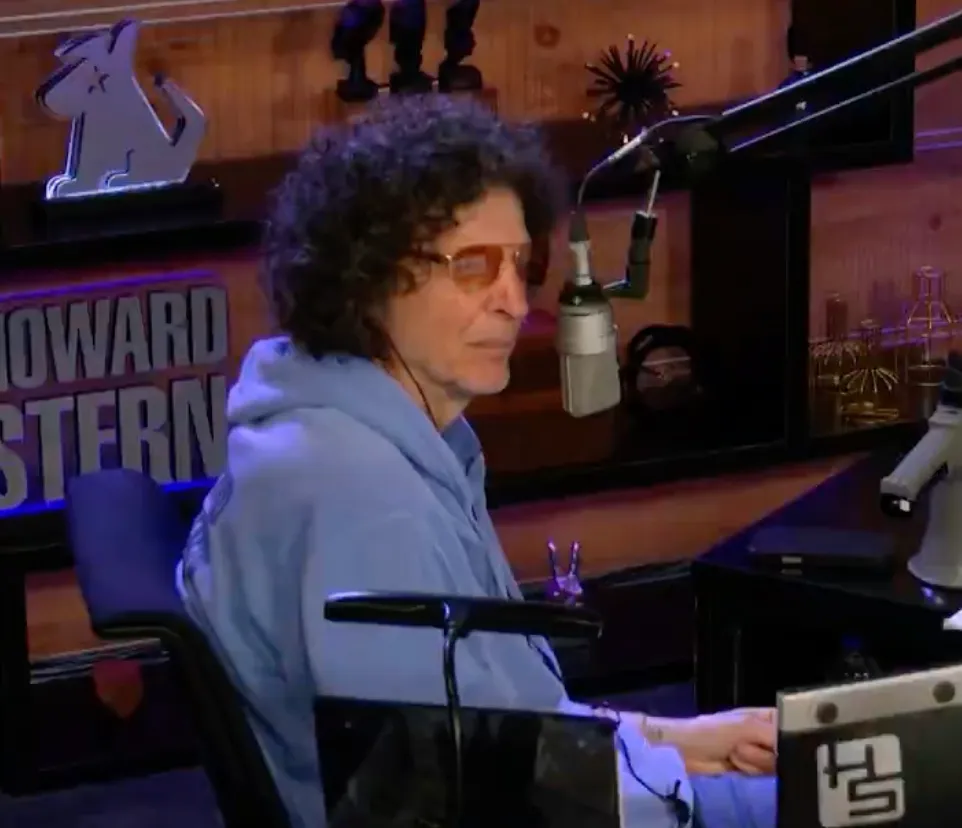 Howard Stern wearing a light blue hoodie and orange sunglasses, looking to the side in his studio.