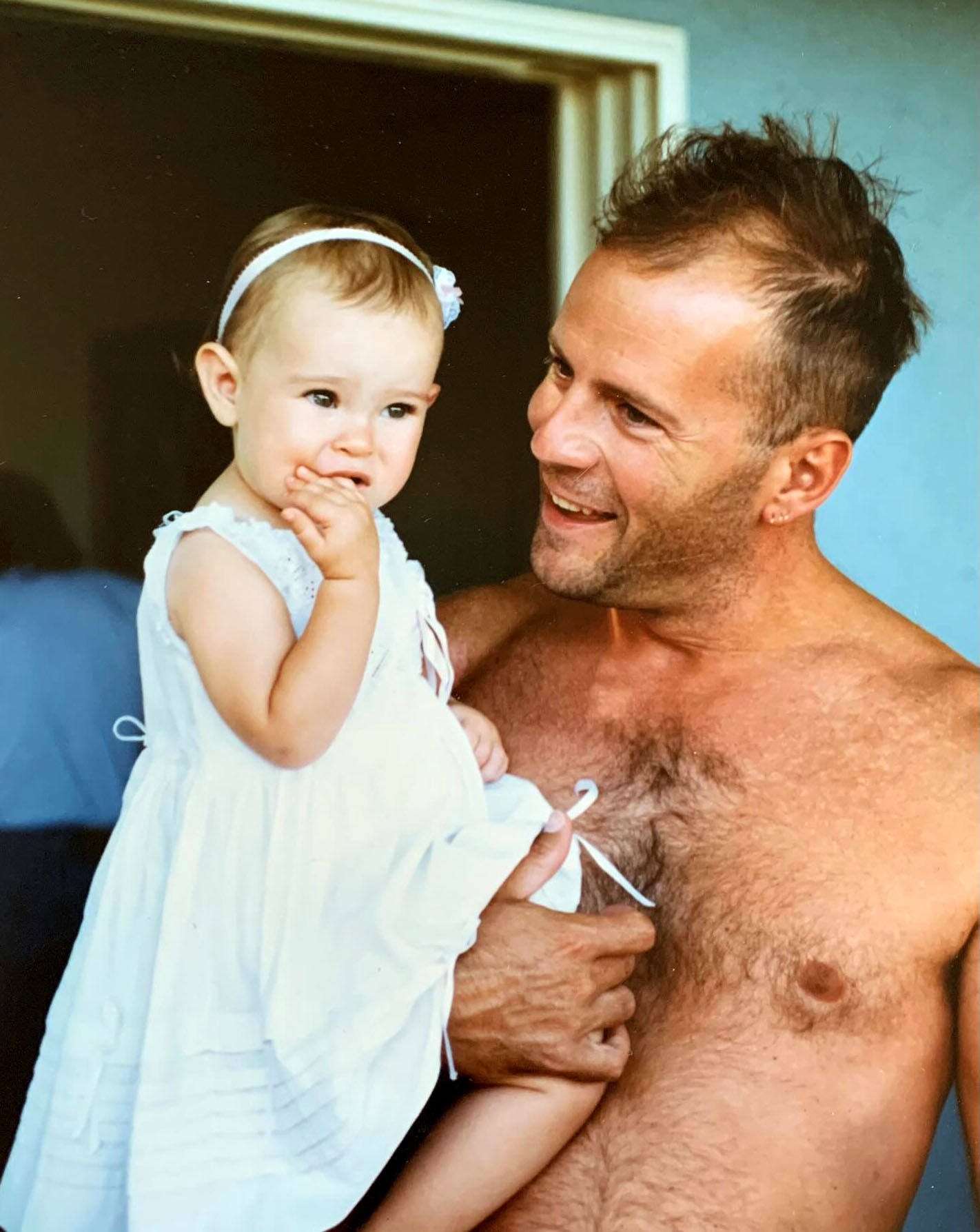 Rumer Willis as a baby in a white dress, held by her father Bruce Willis.