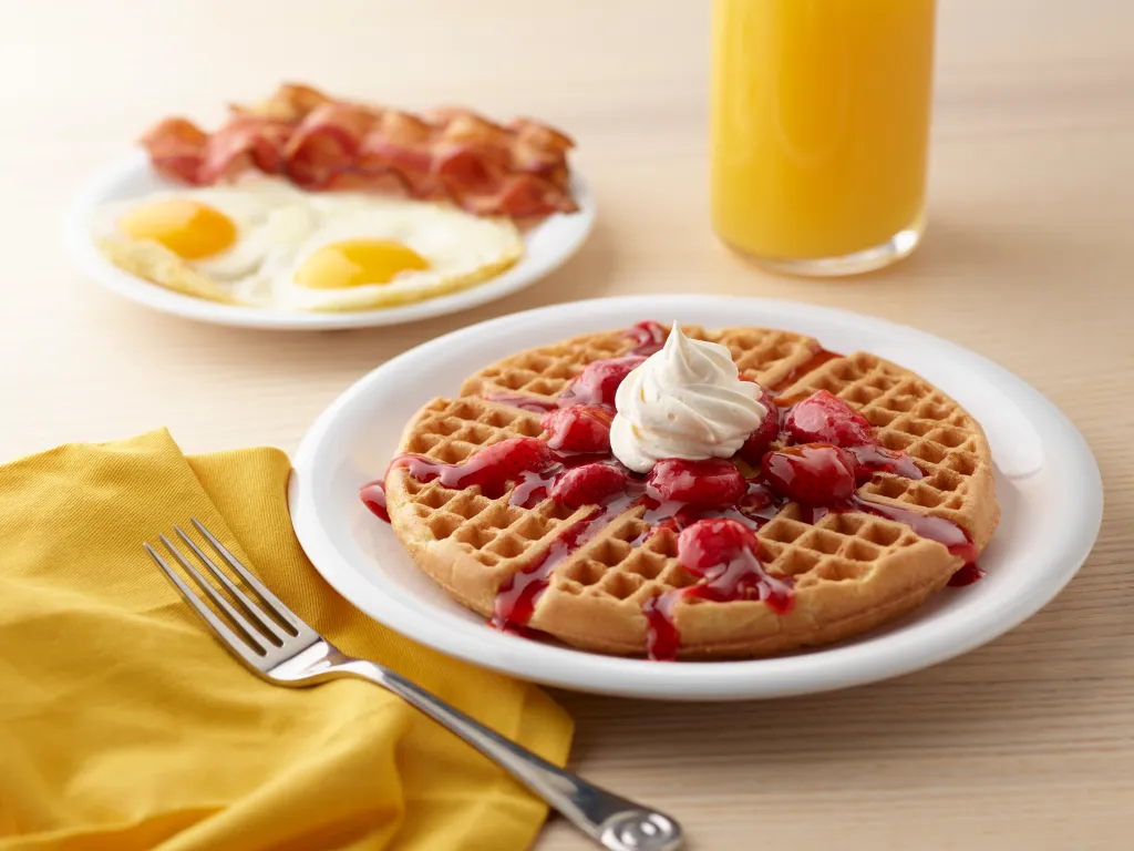 Huddle House Mother's Day Special breakfast featuring a waffle with strawberry topping and whipped cream, a plate of eggs and bacon, and a glass of orange juice.