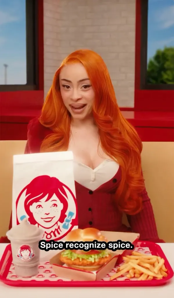 Ice Spice with a Wendy's meal, a Frosty, a crispy chicken sandwich, fries, and a bag, while wearing a red cardigan and white shirt.