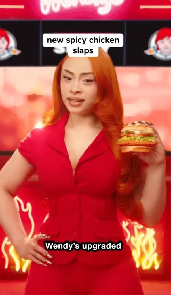 Ice Spice wearing a red outfit and holding a Wendy's spicy chicken sandwich.