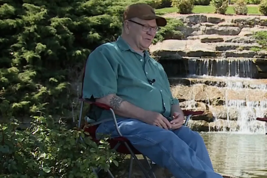 Man wearing a green shirt, jeans, and a baseball cap sitting in a chair outdoors next to a waterfall.
