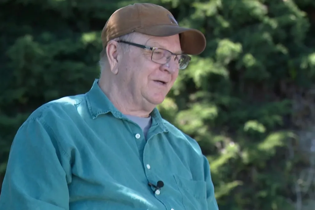 A man in a brown baseball cap, glasses, and a teal shirt smiles.