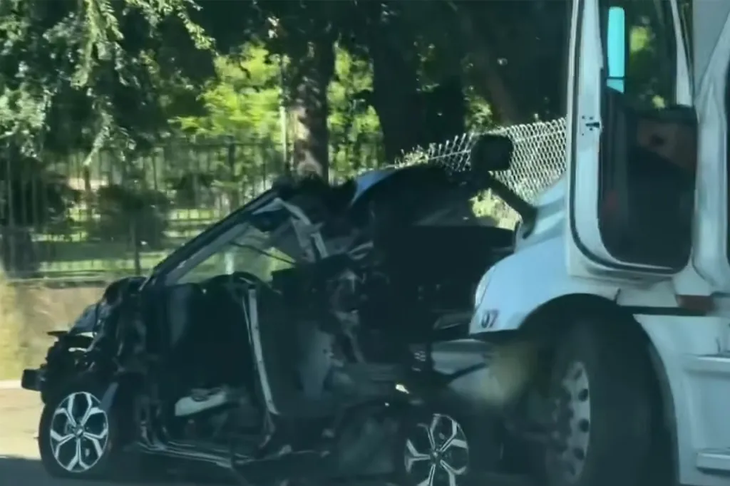 A heavily damaged black car is wedged underneath the front of a white semi-truck.