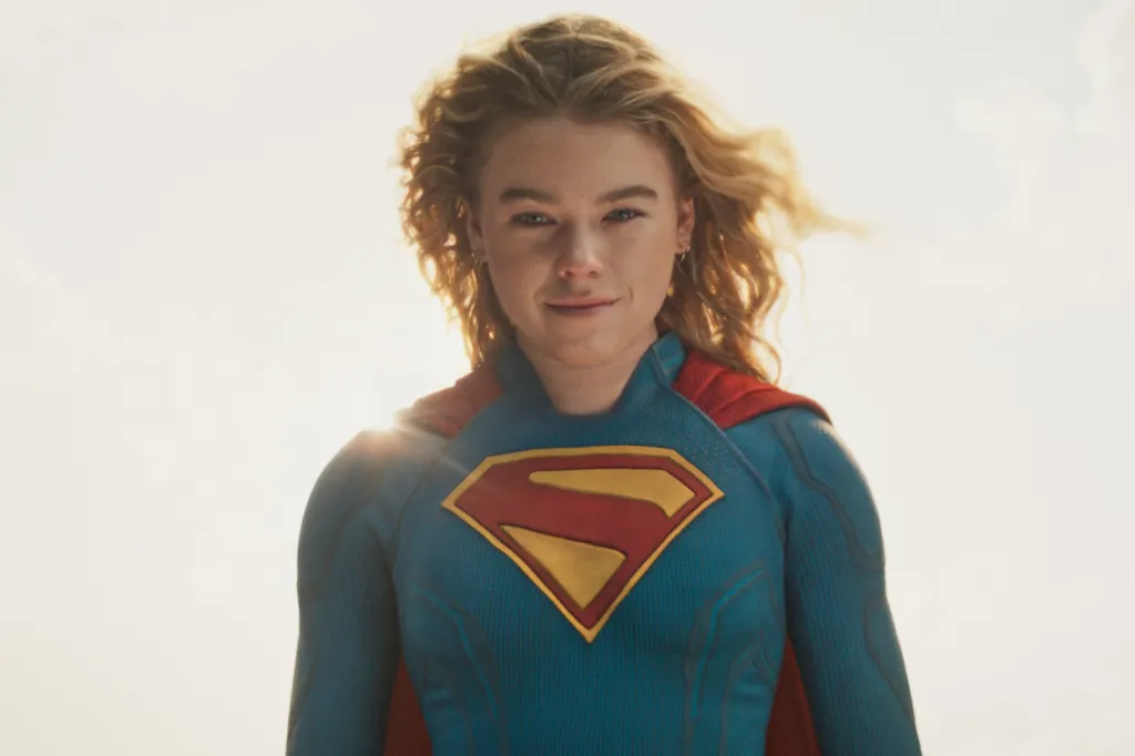 Milly Alcock as Supergirl in a blue suit with red and gold S-shield, with a red cape, smiling.