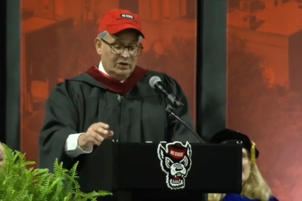 Anil Kochhar, a donor wearing a graduation gown and an NC State baseball cap, speaks at a podium.