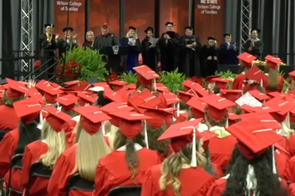 Graduating students in red caps and gowns face a stage where faculty members are applauding.