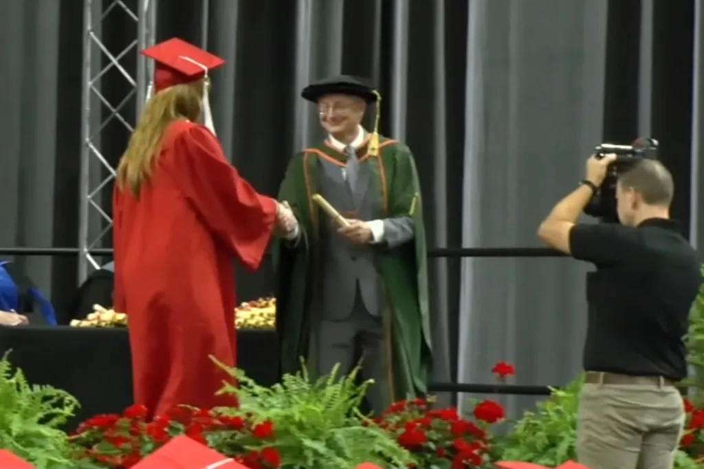 A graduation ceremony where a student in a red gown shakes hands with a man in a green gown, while a photographer captures the moment.