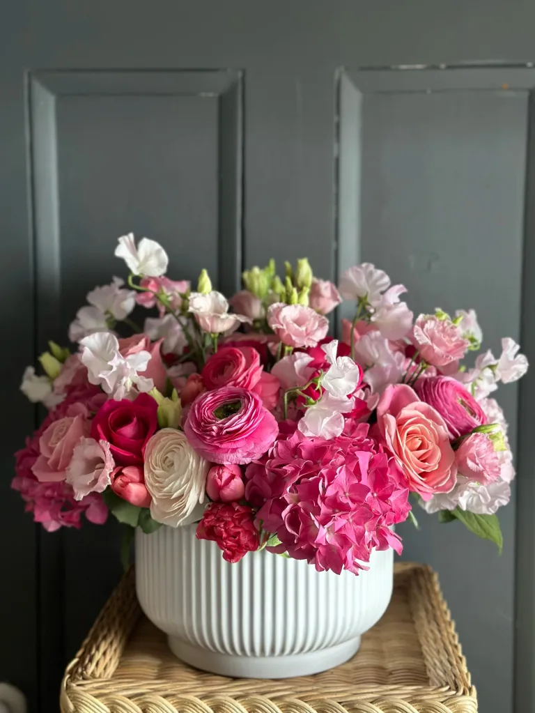 Pink and white flowers in a white fluted vase on a wicker table.