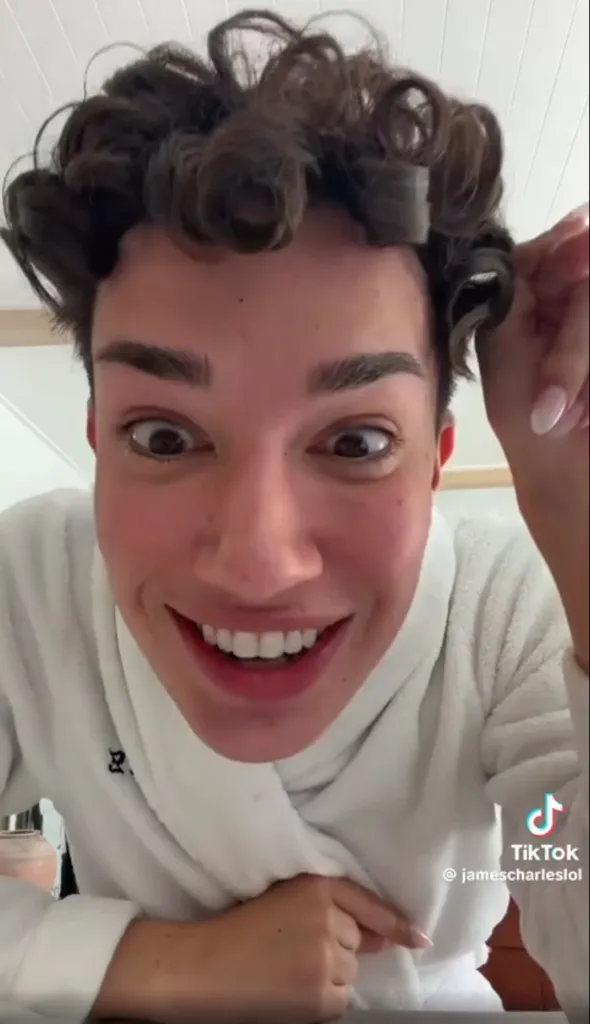 James Charles making a wide-eyed, smiling expression with hair curlers.