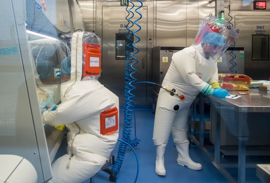 Two workers in full white protective suits and blue head coverings work inside a P4 laboratory.