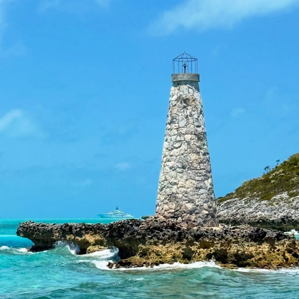 A lighthouse on a rocky coast with a yacht in the background in the Bahamas.