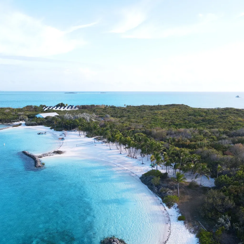 Aerial view of a private island in the Bahamas with white sand beaches, clear blue water, palm trees, and buildings.