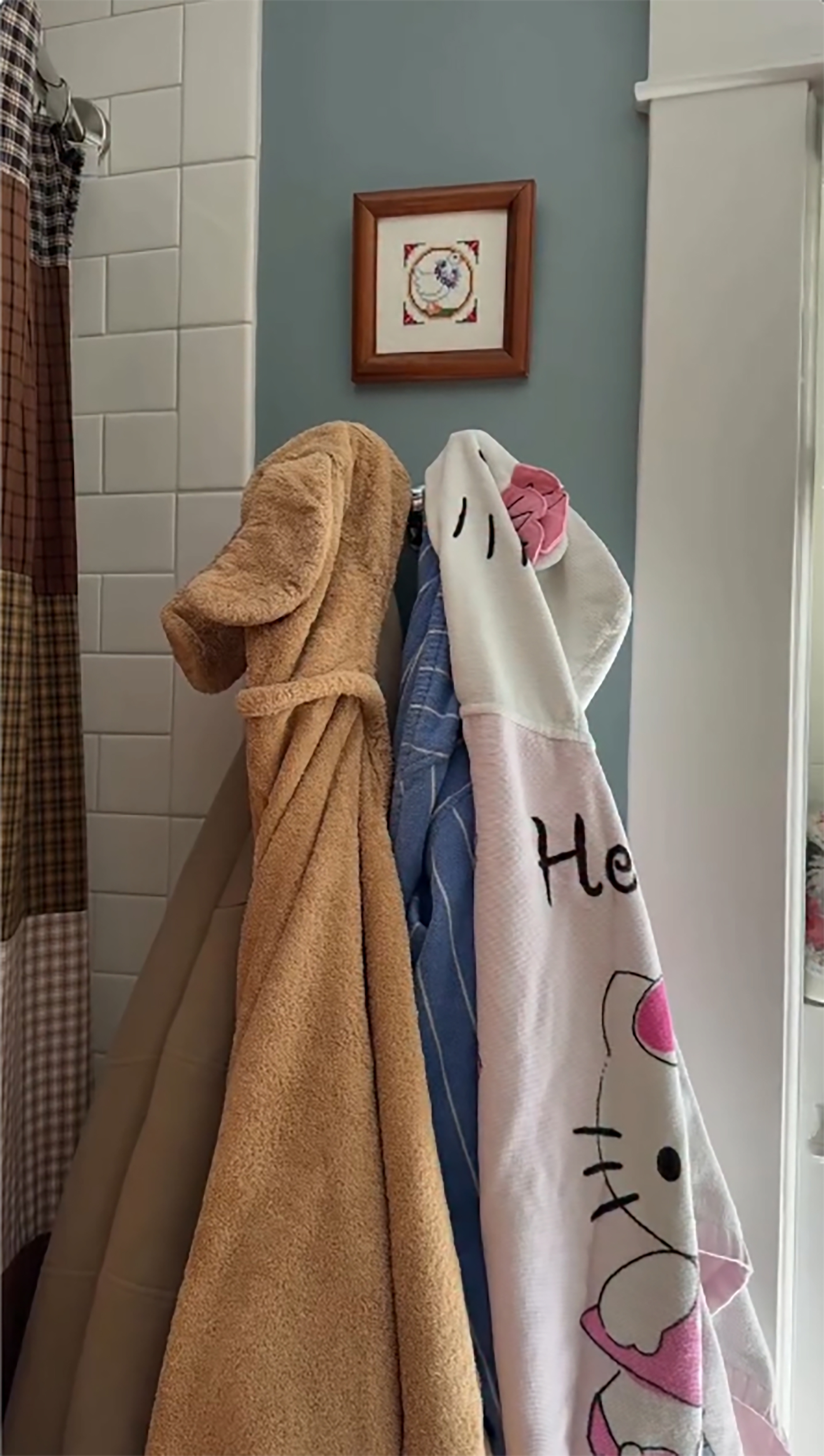 A brown bathrobe and a white Hello Kitty towel hanging on hooks next to a shower with a plaid curtain.