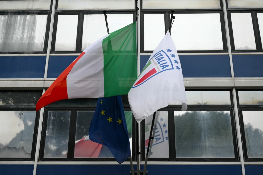 The Italian national flag and the Italian Football Federation (FIGC) flag hang on poles in front of a building with blue walls and large windows.