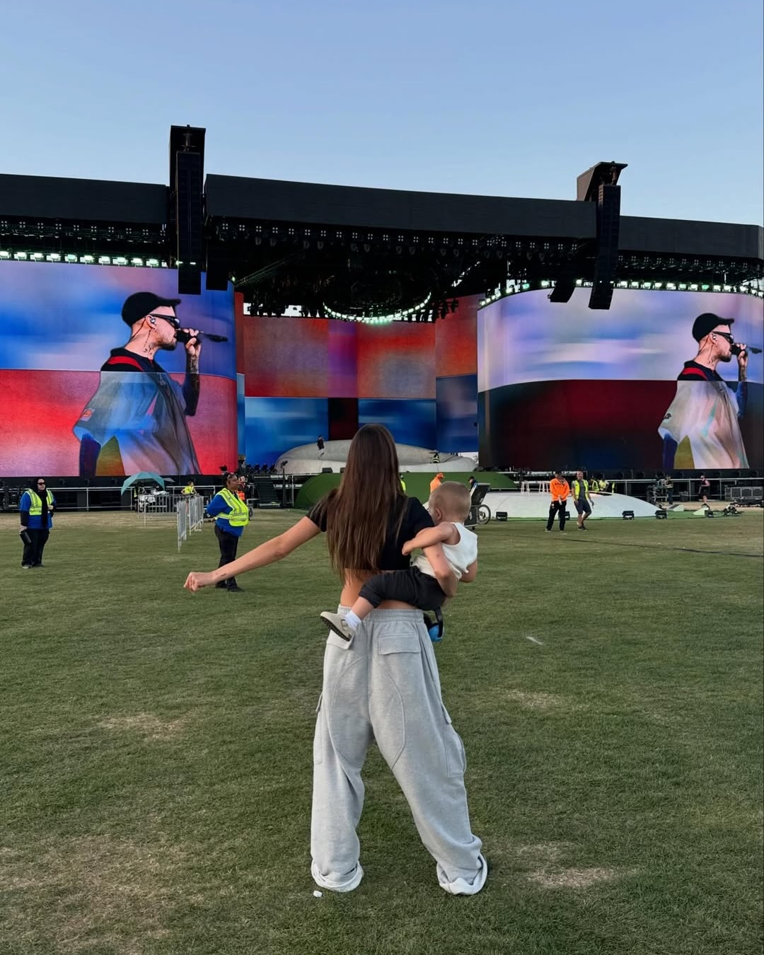 Hailey Bieber holding a baby while watching a performer on large screens at an outdoor concert.
