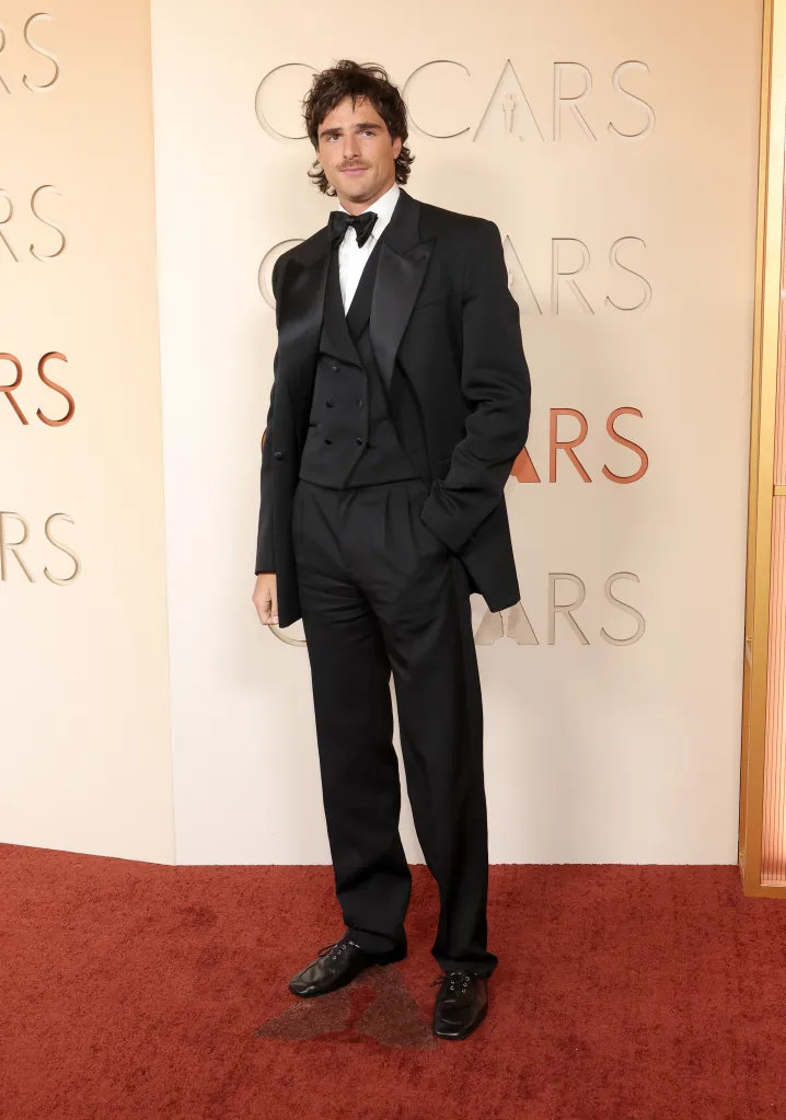 Jacob Elordi on the red carpet at the 98th Oscars, wearing a black tuxedo with a black bow tie and vest.