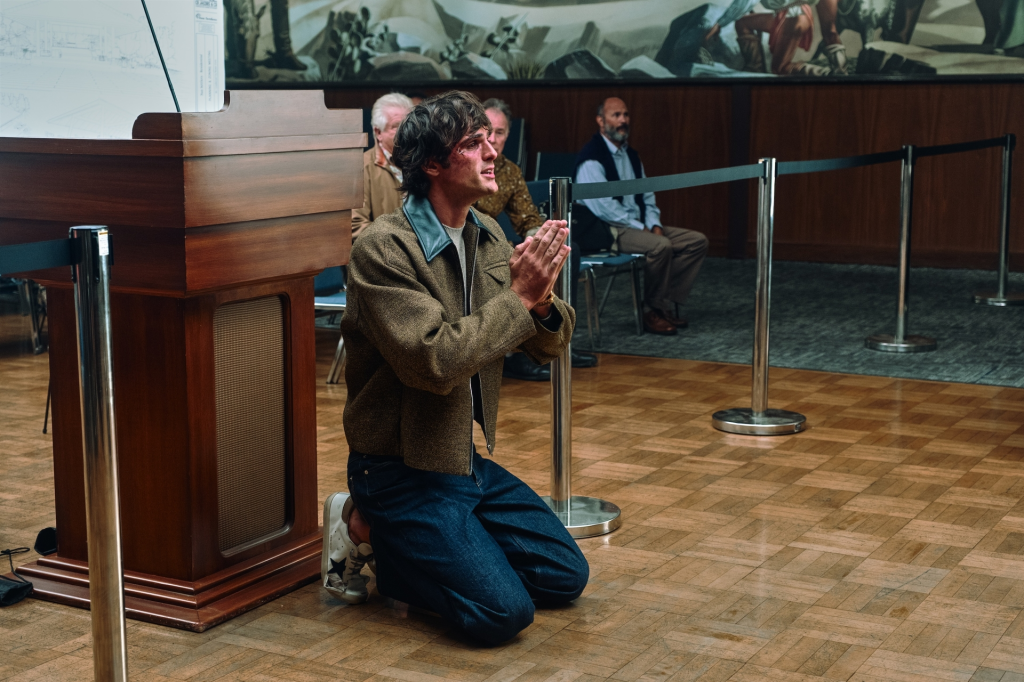 Jacob Elordi kneeling with hands clasped in front of a wooden stand.