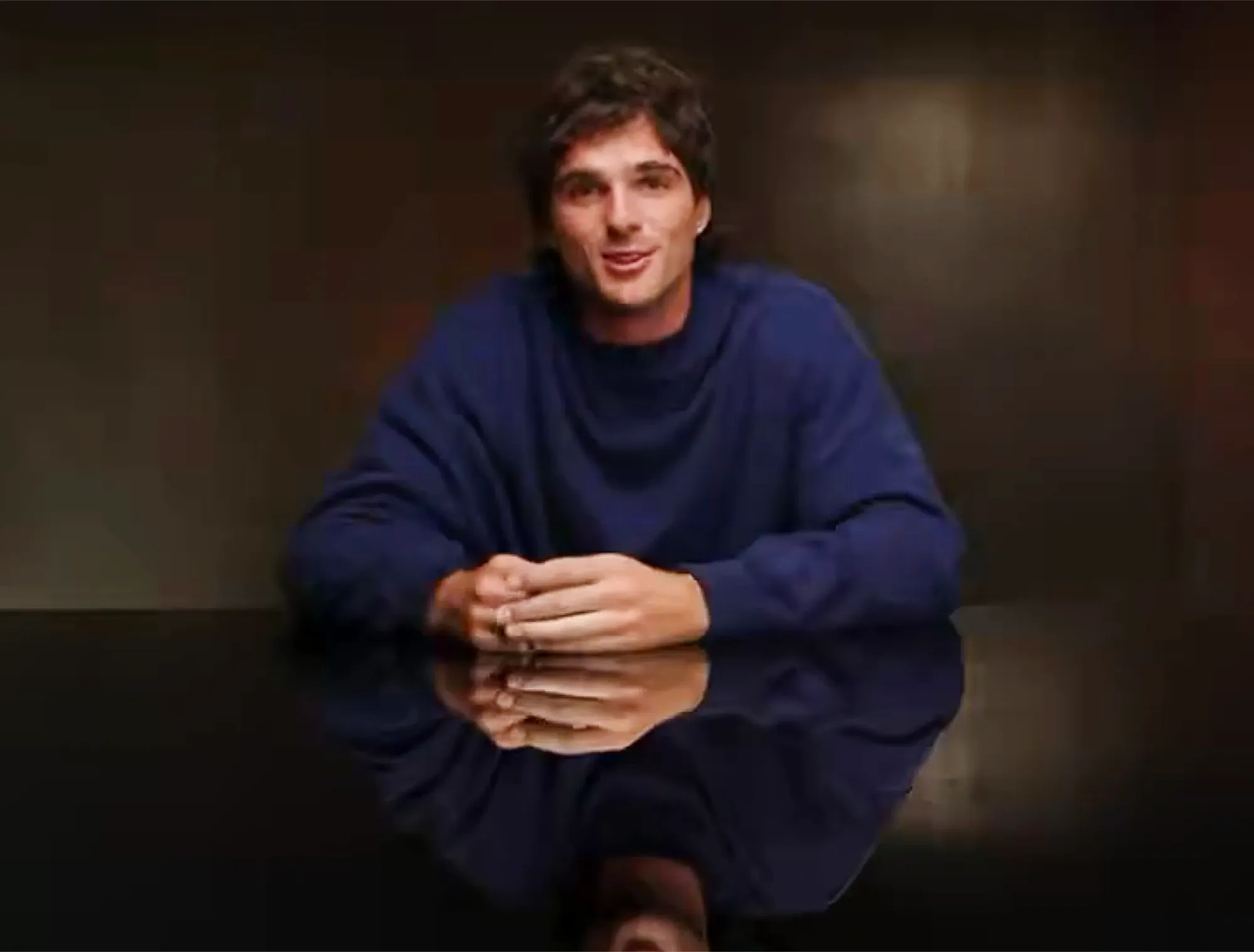 Jacob Elordi wearing a dark blue crew neck sweatshirt, seated with clasped hands on a reflective dark surface.