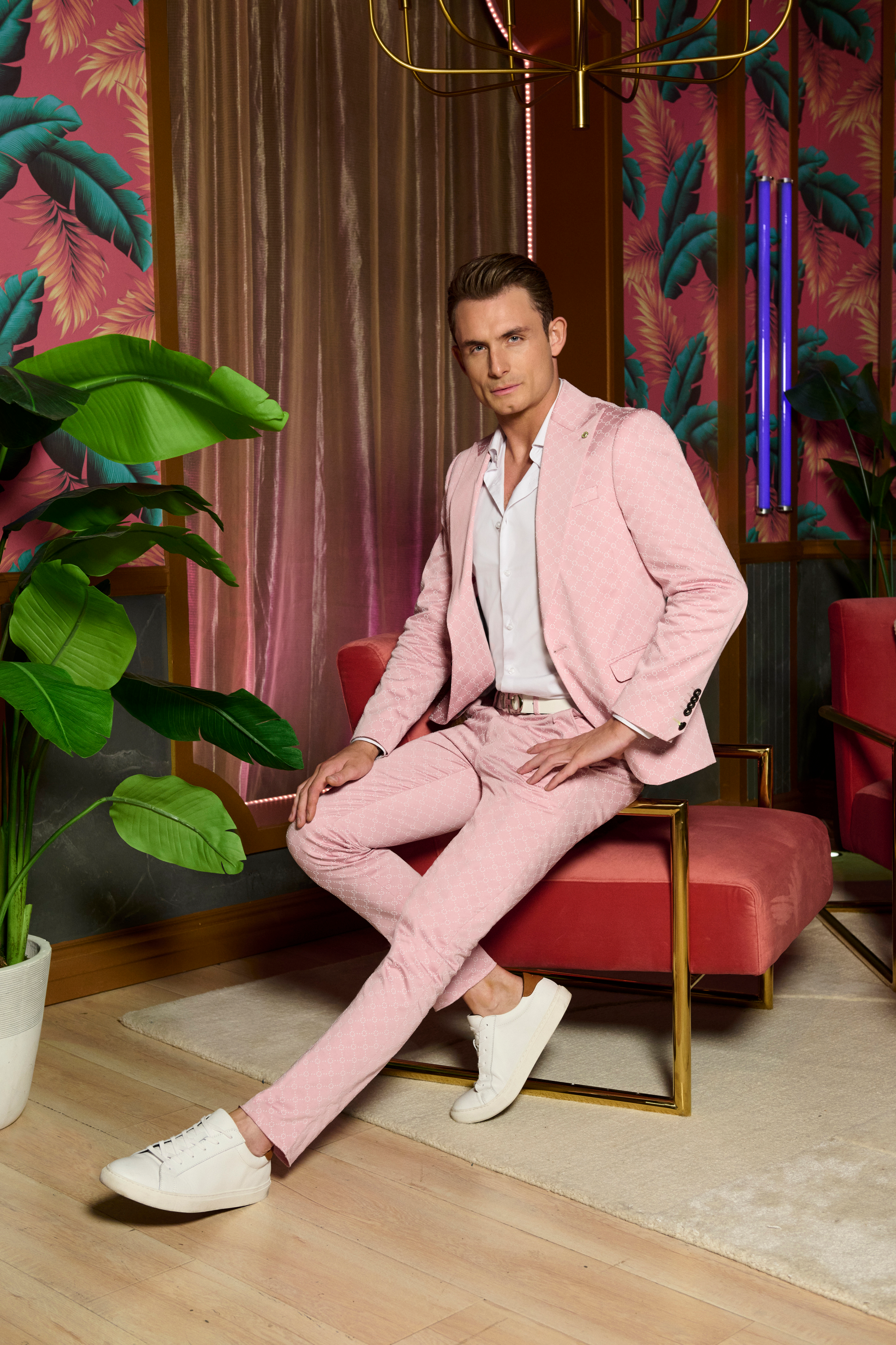 James Kennedy sitting on a red velvet chair, dressed in a pink suit, white shirt, and white sneakers, with a tropical-themed wall and plant in the background.