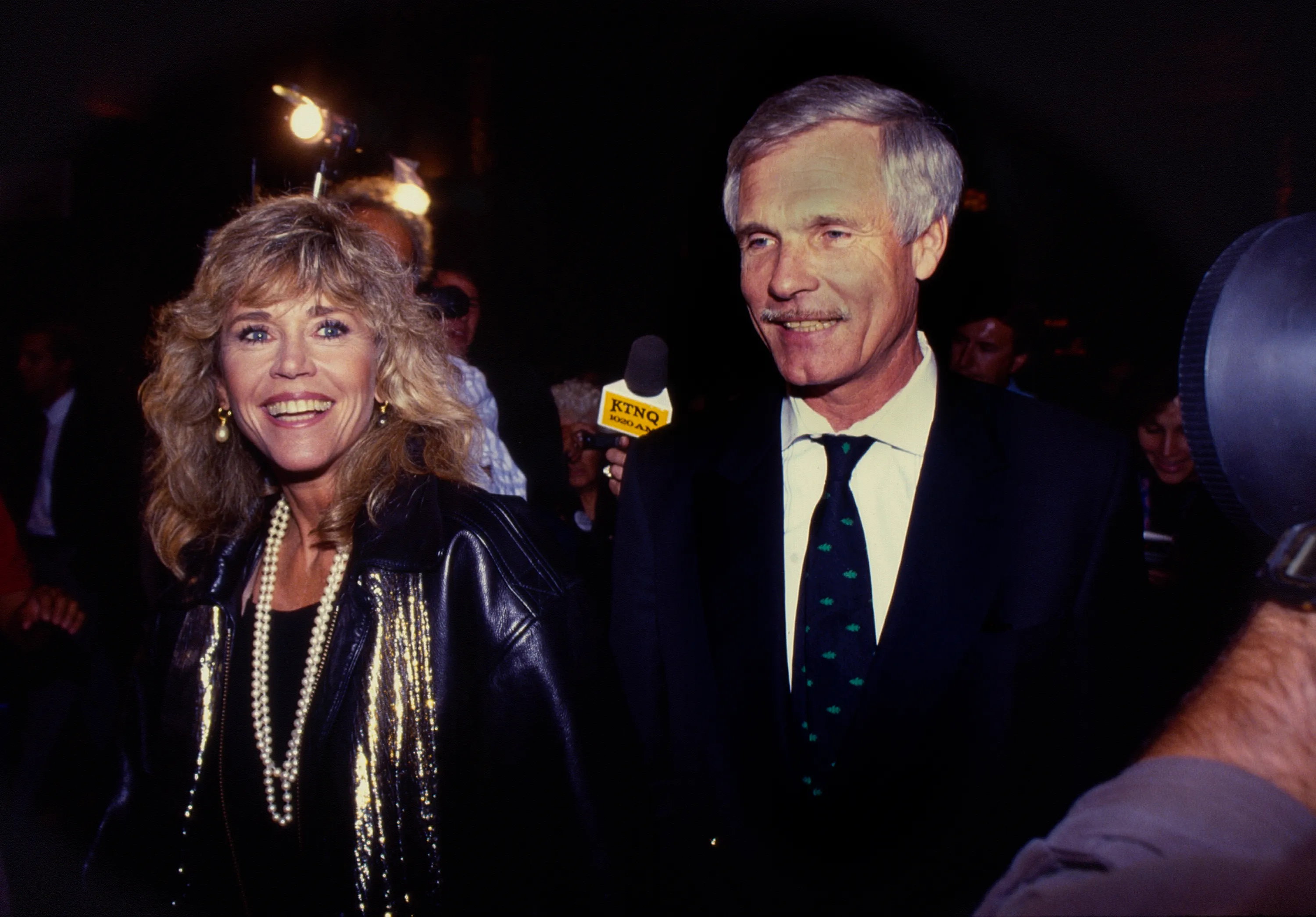 Jane Fonda and Ted Turner smiling in a low-light setting with camera flashes and a microphone visible.