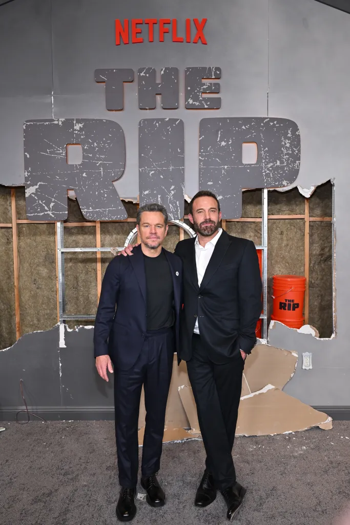 Matt Damon and Ben Affleck standing on a gray carpet in front of a wall with holes showing the insulation and studs, with the Netflix and The Rip logos on the wall.